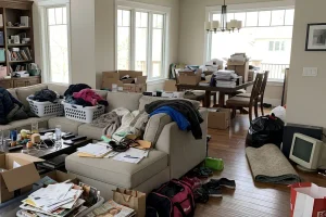 Cluttered living room and dining area filled with boxes, clothing, household items, and belongings awaiting a home cleanout 